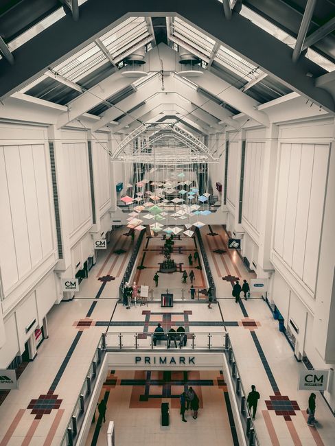 A wide aerial view of the spacious shopping mall interior on a bright day, showcasing polished ceramic tile floors reflecting the overhead lighting. The corridor features large glass storefronts, incl